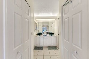 A hallway with white doors leading to a bathroom with a double sink and a large mirror. - Coral Princess Waterfront Pool & Hot Tub (Cape Coral)