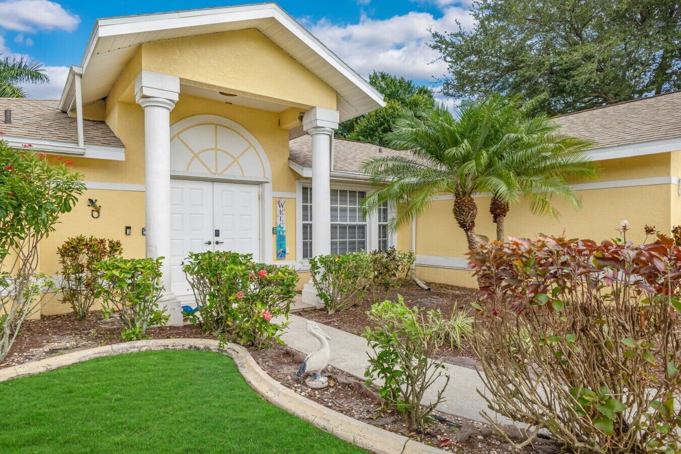 A yellow building with a white door, surrounded by a well-maintained garden with various plants and a palm tree.