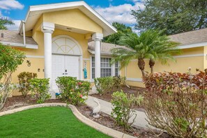 Um edifício amarelo com porta branca, rodeado por um jardim bem cuidado com diversas plantas e uma palmeira.