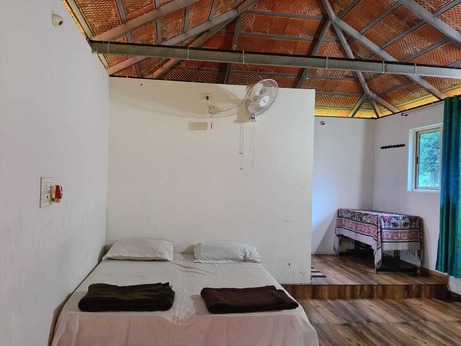 A single bed with two pillows, a ceiling fan, a small table with a patterned tablecloth, and a window with curtains.