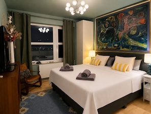 A hotel room with a bed, a desk, a chair, a window, a painting, and a chandelier.