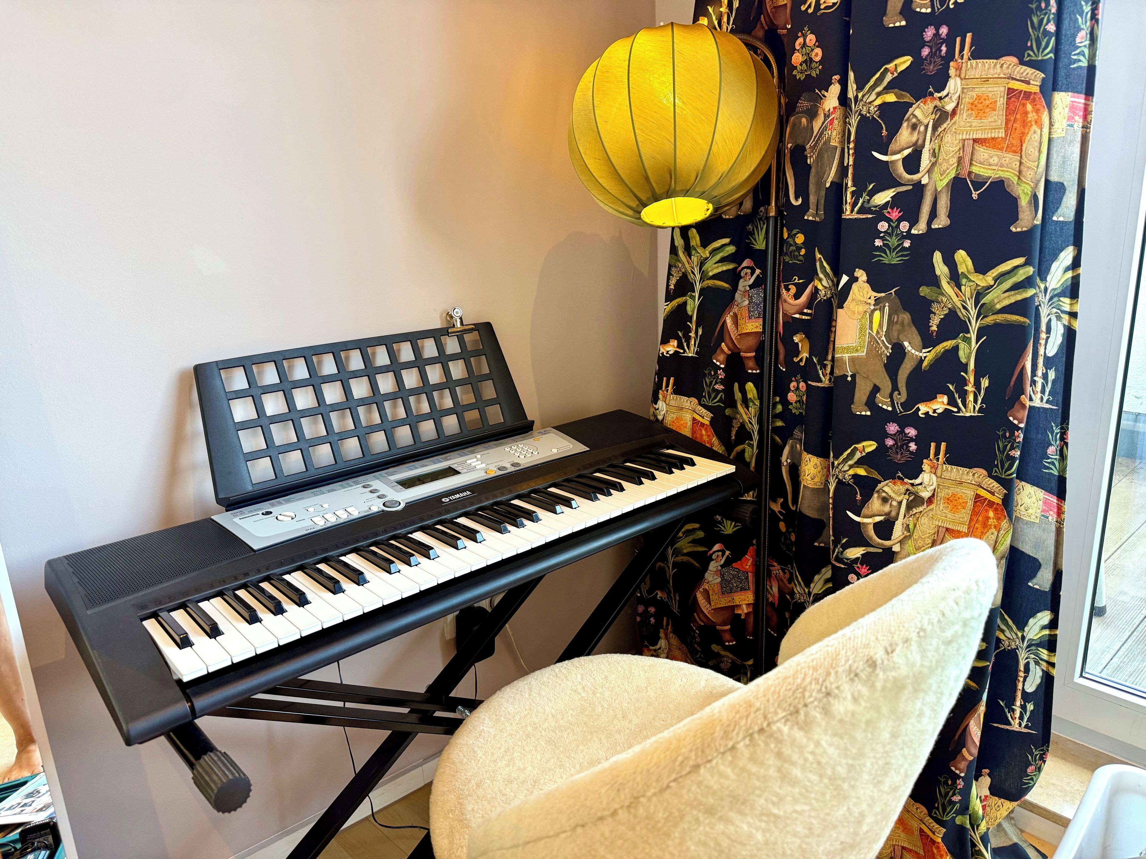 A keyboard with a stand, a chair, a lamp, and a curtain with an elephant print.