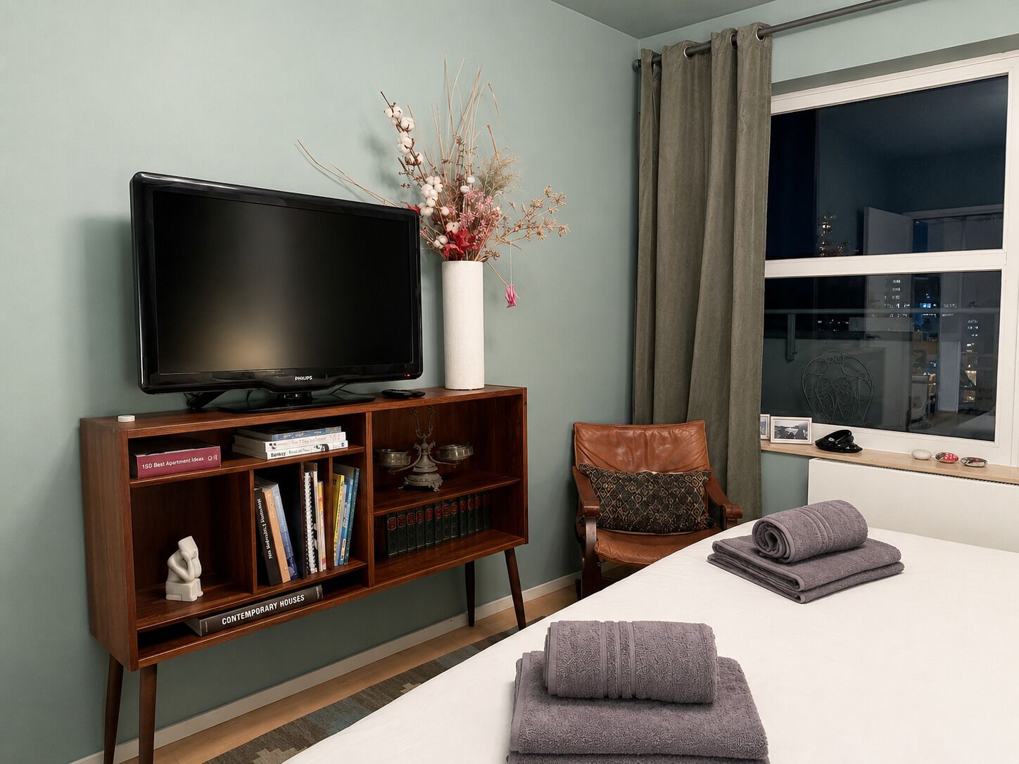 A hotel room with a television on a wooden stand, a bookshelf with books, a decorative vase with dried flowers, a brown leather chair, and a bed with folded towels.