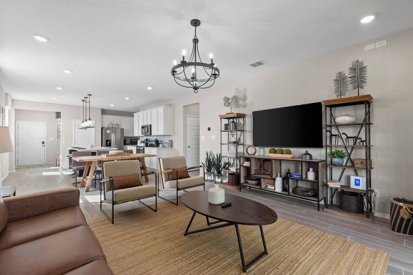 Una sala moderna con sofá, mesa de centro y comedor con sillas. Se observa una cocina al fondo, un televisor montado en la pared y una estantería con diversos objetos.