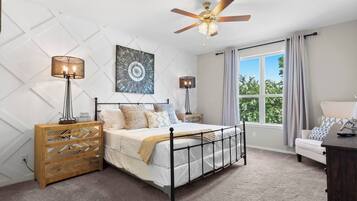 A bedroom with a bed, a wooden dresser, a ceiling fan, a window with curtains, and a view of greenery outside.