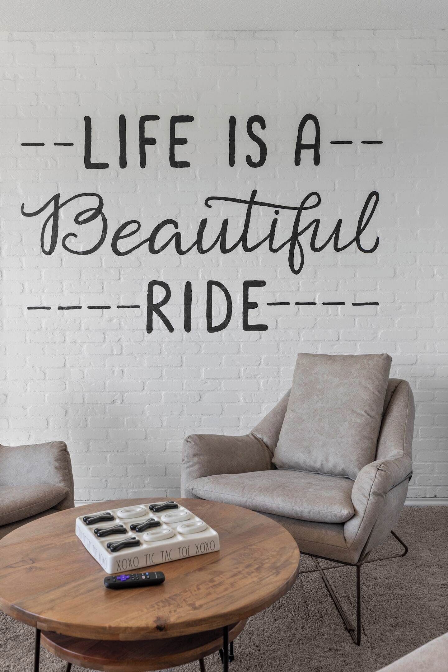 A room with a white brick wall, a motivational quote, a beige armchair, a round wooden coffee table, and a remote control.