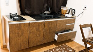 A kitchenette with a gas stove, a sink, and a coffee maker. There is a wooden cabinet with drawers and a wooden chair. A coffee-themed rug is on the floor.