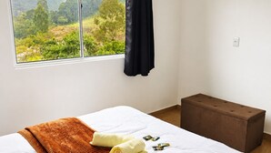 A hotel room with a bed, a window with a view of greenery, a brown ottoman, and a black curtain.