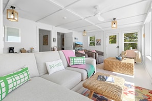 A spacious living room with a large sofa, a coffee table, and a television. There are doors leading to other rooms, and a window with a view of trees. - Camp Avaliz - Ocean-view for 2 with a queen bedroom, kitchenette, Wi-Fi and AC (Gouldsboro)