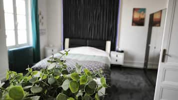 Um quarto com cama, mesa de cabeceira, cortinas na janela, porta e uma planta.