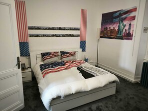 A bedroom with a bed, a nightstand, a lamp, and a wall with a cityscape painting.
