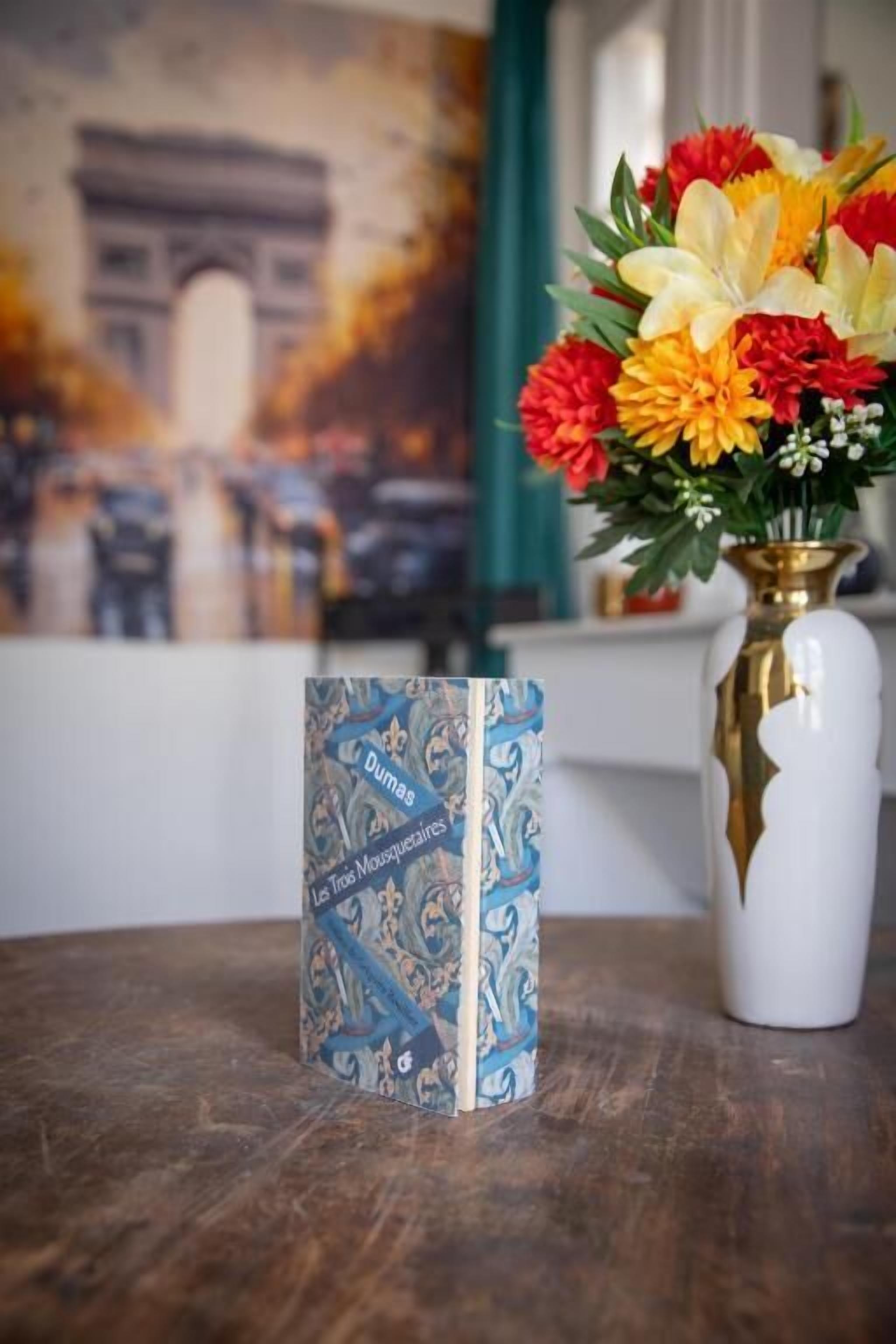 Un libro titulado 'Dumas Les Trois Mousquetaires' sobre una mesa con un jarrón de flores.