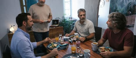 Un grupo de personas cenando en una mesa de madera en una habitación con un espejo grande, una planta y un mural tropical.