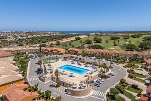 A residential area with a swimming pool surrounded by lounge chairs and umbrellas, a road with cars, and a view of the ocean. - Boavista Lakeview | Golf & Pool Retreat (Lagos)