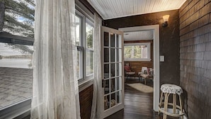 Una habitación con piso de madera, una cortina blanca, un taburete y una puerta que da a otra habitación.