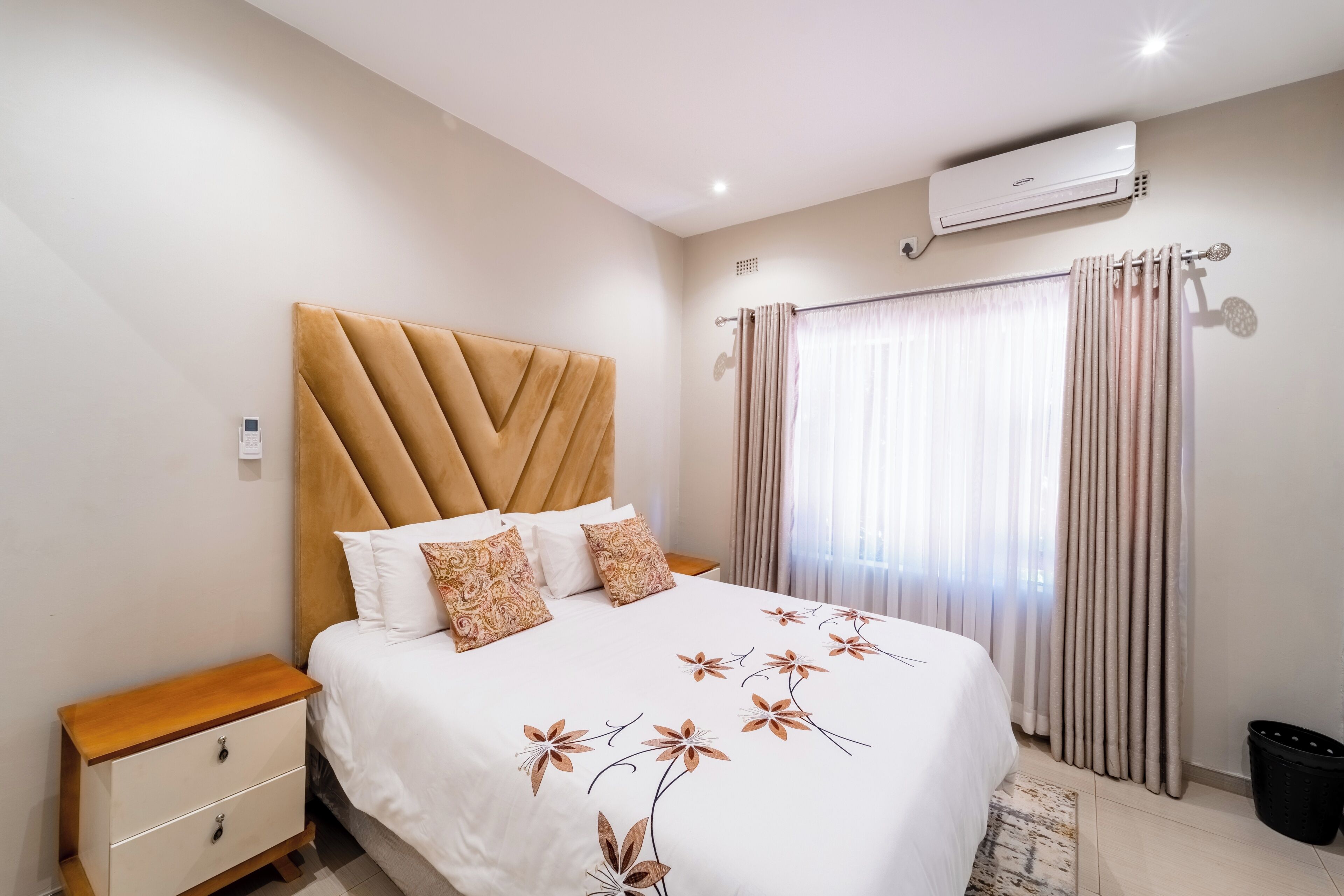 A hotel room with a bed, a nightstand, a wall-mounted air conditioner, and a window with curtains.