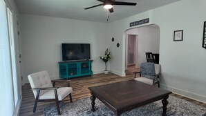 Een woonkamer met een televisie, een entertainmentmeubel in blauwgroen, twee stoelen, een salontafel, een plafondventilator en een bordje met de tekst 'wees onze gast'.