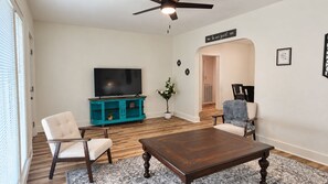 A living room with a wooden floor, a dark wooden coffee table, a teal entertainment unit, a flat-screen TV, and two chairs. - Cozy 2 Bedroom Monthly (Hot Springs)