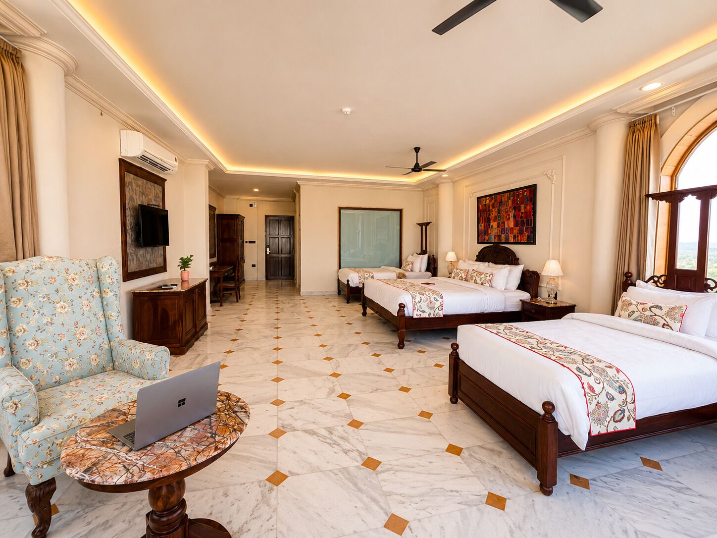 A hotel room with two beds, a sofa, a desk with a laptop, a television, and a large window.
