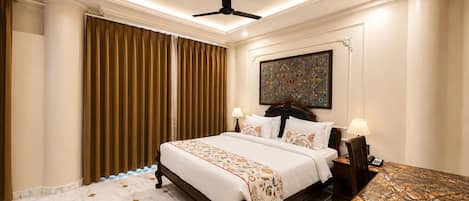 A hotel room with a bed, a desk, a ceiling fan, and a painting on the wall.