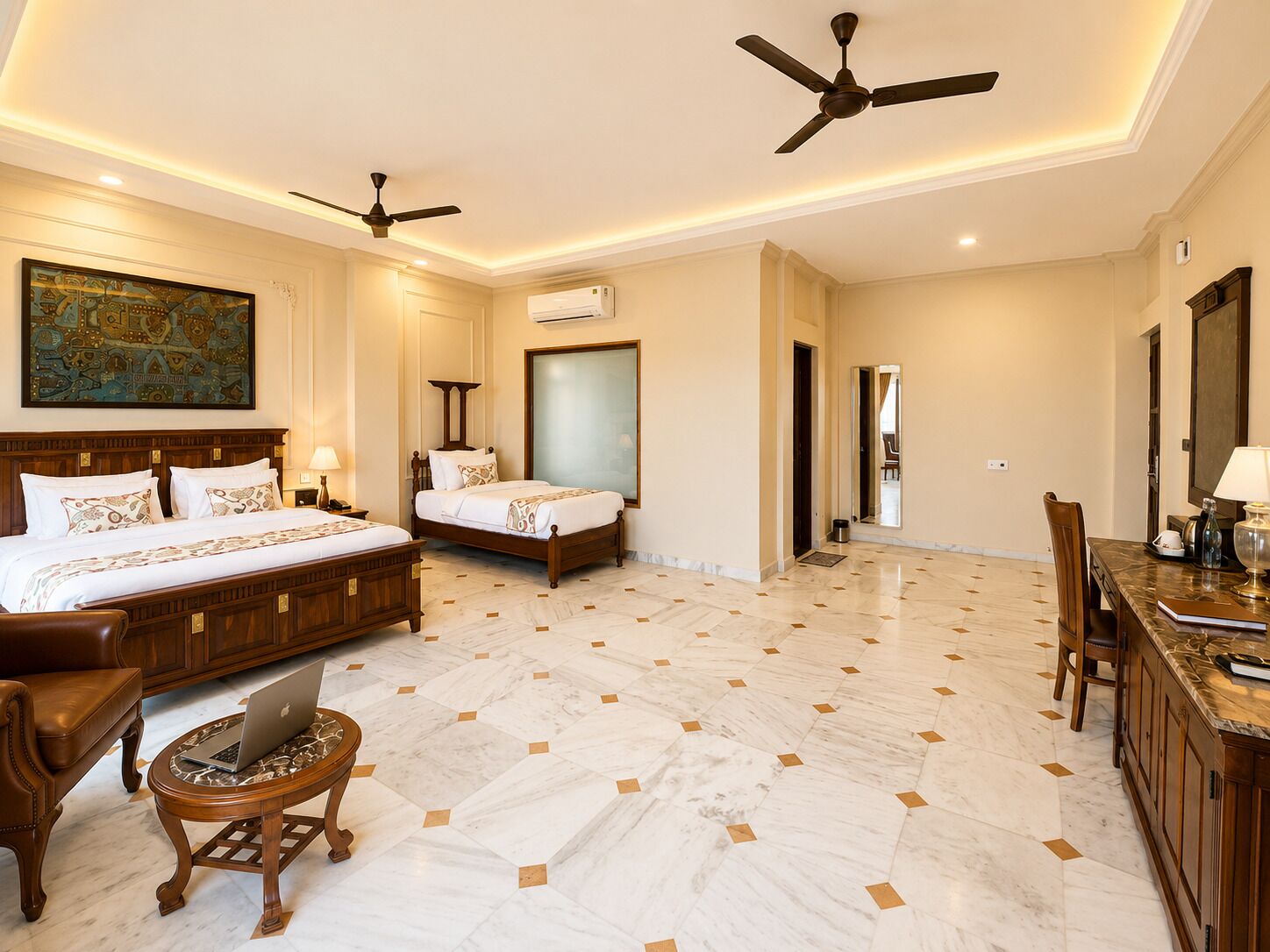 A hotel room with two beds, a desk with a laptop, a dining area, and a ceiling fan.