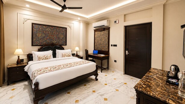 A hotel room with a bed, bedside tables, a desk with a laptop, a ceiling fan, and a large mirror. - Barliyas Heritage Boutique Hotel (Udaipur)