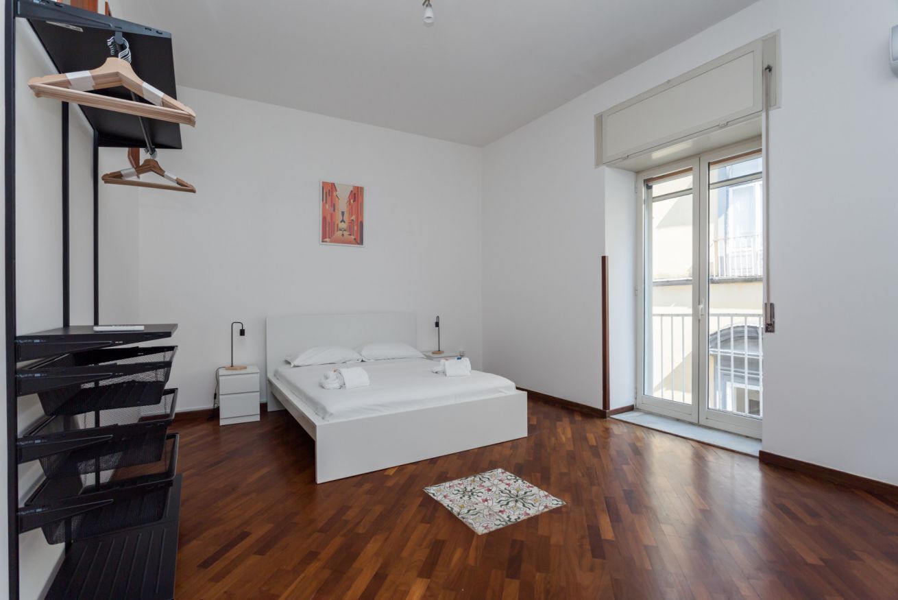 Een slaapkamer met een bed, bureau, stoel, nachtkastje, kledingrek en een raam.