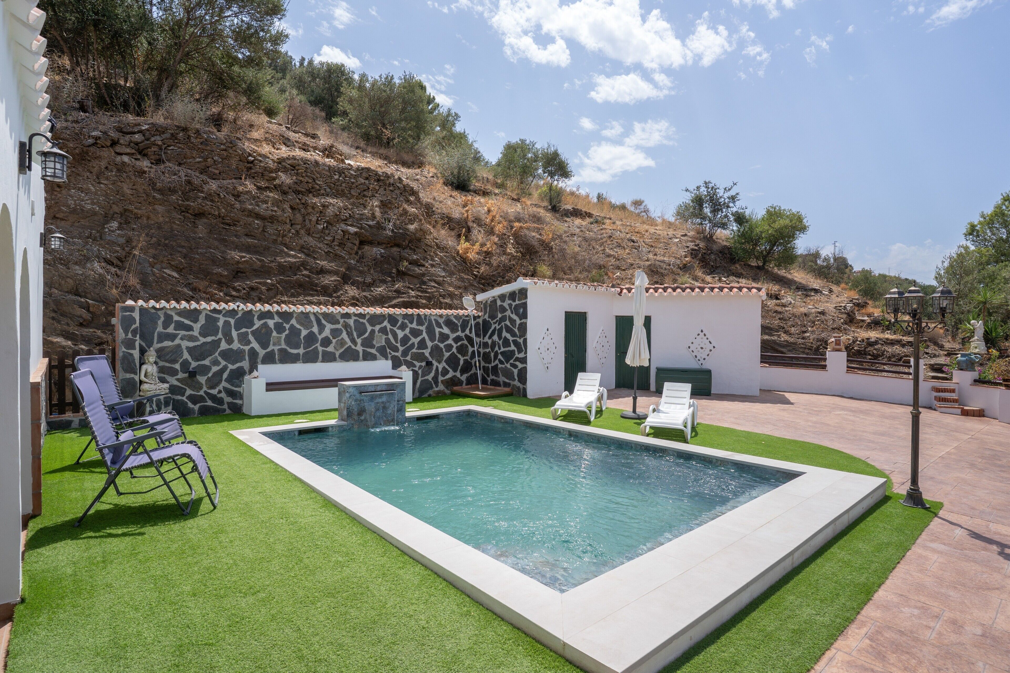 A swimming pool with a stone wall and a small building, surrounded by artificial grass and outdoor furniture.