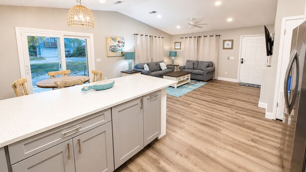Una cocina moderna con una isla grande, un comedor con mesa y sillas, una sala de estar con sofá y alfombra, y vistas al exterior por una puerta corrediza.
