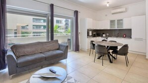 A modern apartment with a grey sofa, a round dining table with chairs, a balcony with curtains, and a kitchen with white cabinets. - Modern 1BR in Fort Cambridge, Sliema | AC & WIFI (Sliema)