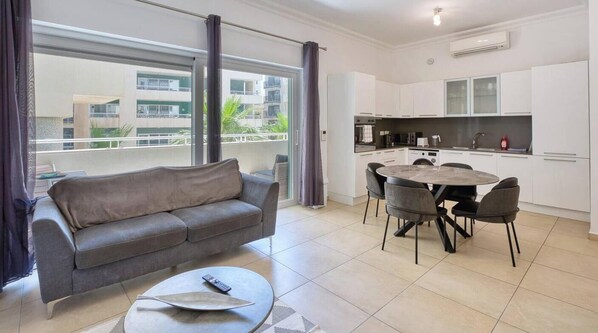 Un apartament modern amb un sofà gris, una taula de dinar rodona amb cadira, un balcó amb cortines i una cuina amb armaris blancs.