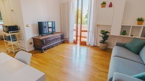 Ein modernes Wohnzimmer mit Holzboden, einem grauen Sofa, einem Fernseher auf einer Holzkommode und einem Balkon mit weißen Vorhängen.
