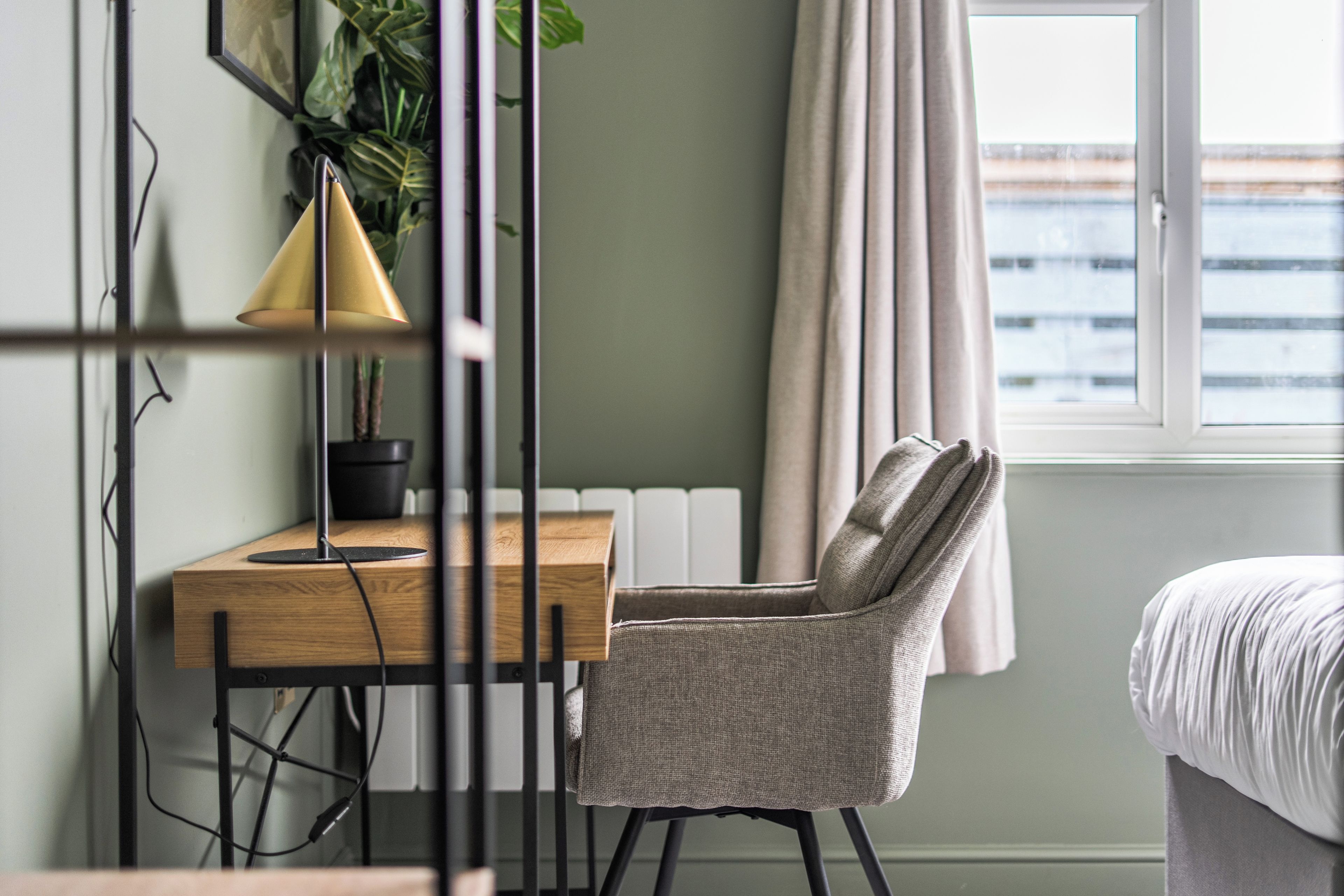 A room with a wooden desk, a chair, a lamp, a potted plant, a radiator, a window with curtains, and a bed.