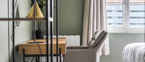 A room with a wooden desk, a chair, a lamp, a potted plant, a radiator, a window with curtains, and a bed.