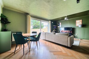 Ein modernes Wohnzimmer mit einer Couch, einem Esstisch, Stühlen, einem Fernseher und einer Topfpflanze.