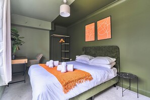 Ein ordentlich eingerichtetes Schlafzimmer mit einem Bett, einem Schreibtisch, einem Stuhl, einer hängenden Pflanze und zwei abstrakten Kunstwerken an der Wand.