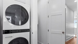A laundry room with stacked washers and dryers, a white door, and a wooden floor. - Luxury Oceanfront, Pool, Elevator, Volleyball (Oak Island)