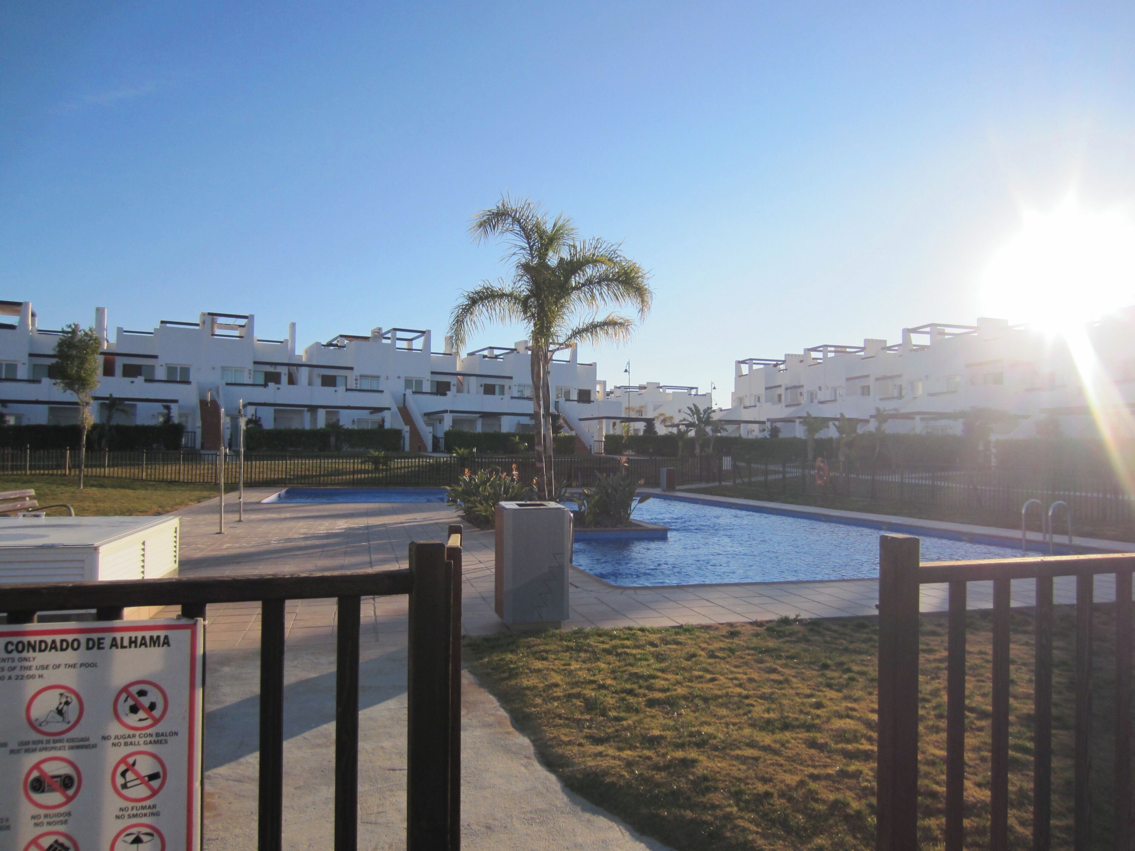 A swimming pool area with a sign indicating pool rules, surrounded by modern buildings and a palm tree.