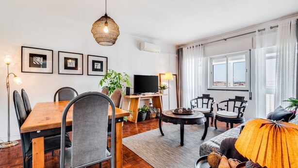 Una sala de estar moderna con una mesa de comedor de madera, sillas tapizadas en gris, un televisor de pantalla plana y un amplio ventanal con cortinas.