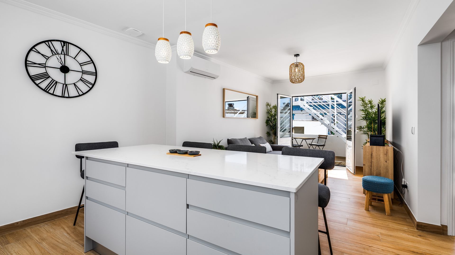 A modern kitchen with a central island, pendant lights, a wall clock, and a view of the outside.