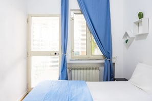 Une chambre à coucher avec un lit, une fenêtre avec des stores, un radiateur et une petite plante posée sur une étagère.