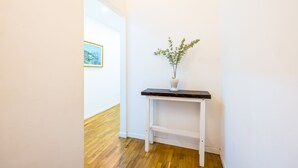 Un pasillo con pared blanca, suelo de madera, una pequeña mesa blanca con un florero de follaje verde y un cuadro en la pared.