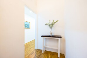 Ein Flur mit weißer Wand, Holzboden, einem kleinen weißen Tisch mit einer Vase mit Grünpflanzen und einem gerahmten Bild an der Wand.