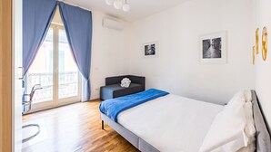 Un dormitorio con cama, sofá, balcón con sillón y dos cuadros en la pared.