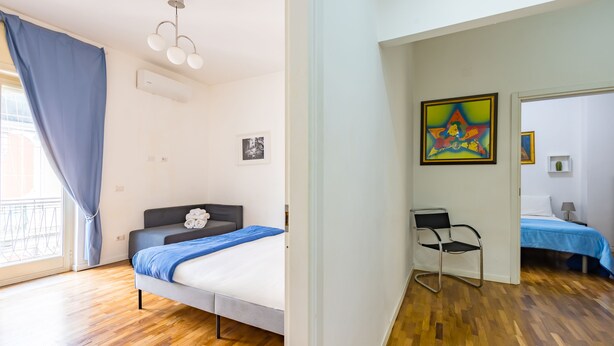 Un dormitorio con cama, una silla, un cuadro y una ventana con cortinas.