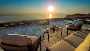 Um terraço com móveis de vime, uma mesa com uma bebida e vista para o pôr do sol.