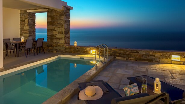 Área à beira da piscina com paredes de pedra, uma mesa com cadeiras e vista para o mar ao entardecer.