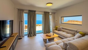 A modern living room with a sofa, a coffee table, and a television. There are large windows with curtains, a view of the sea, and a stone wall.