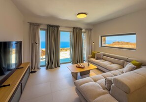 Un salon moderne avec un canapé, une table basse et un téléviseur. De grandes fenêtres, ornées de rideaux, offrent une vue sur la mer, et un mur en pierre ajoute une touche d’élégance.