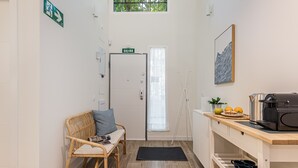 Un pasillo estrecho con una silla de mimbre, un banco, una puerta con un letrero, un ventanal, una lámpara en la pared y una zona de cocina con un hervidor y fruta.